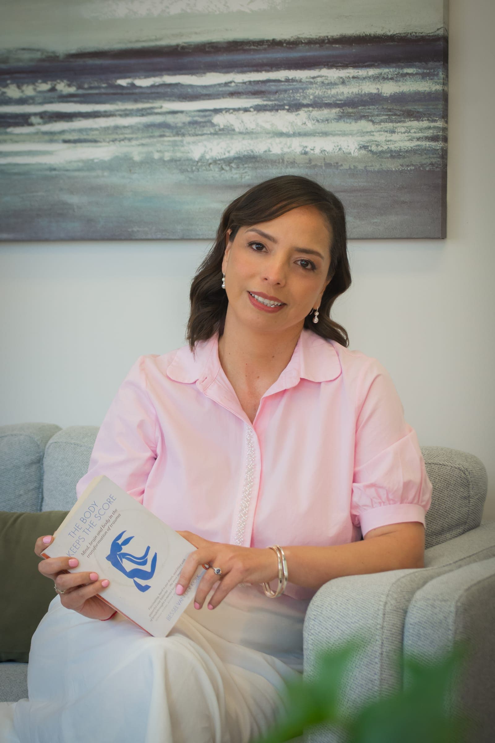 Alpha Ponce smiling on a sofa in her therapy room, holding The Body Keeps the Score
