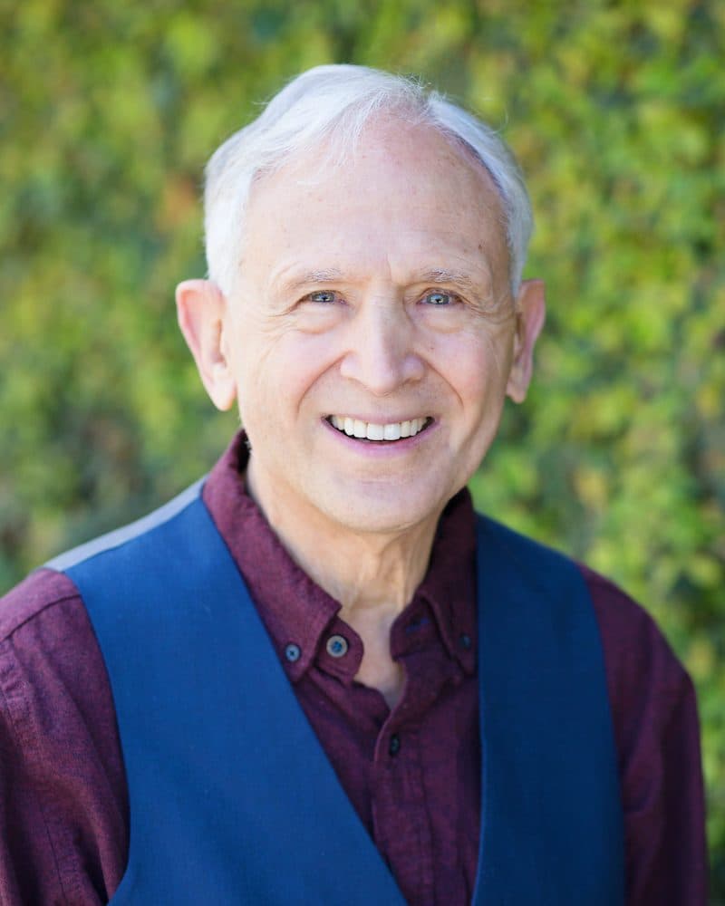 Retrato del Dr. Peter A. Levine, sonriendo, con camisa granate y chaleco azul.