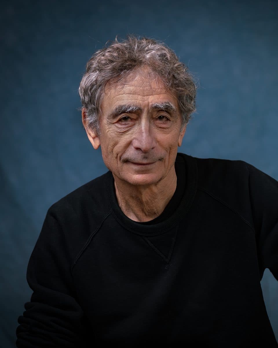 Portrait of Dr Gabor Maté, smiling, in a black sweater against a blue backdrop.